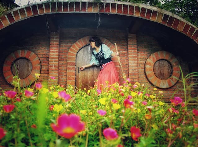 Seorang berdiri di depan spot foto rumah hobbit dengan bunga disekelilingnya di Narmada Botanic Garden