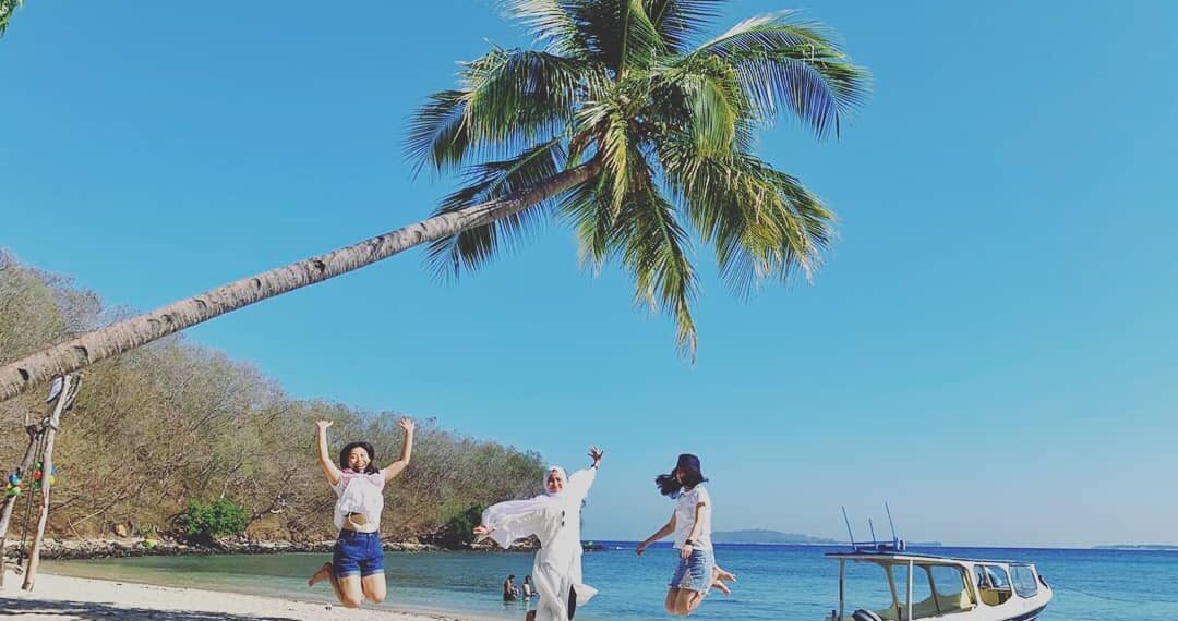 Wisatawan loncat di bawah pohon kelapa di pinggir pantai yang biru