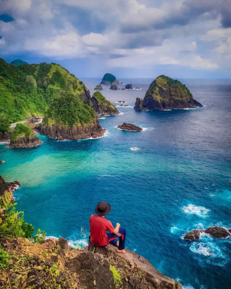 Wisatawan duduk di atas bukit menghadap ke lautan dan Pulau Delapan