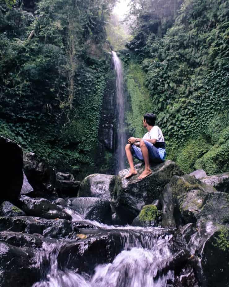 Wisatawan duduk di atas batuan alam memandang ke Air Terjun Titian Batu Kawangan