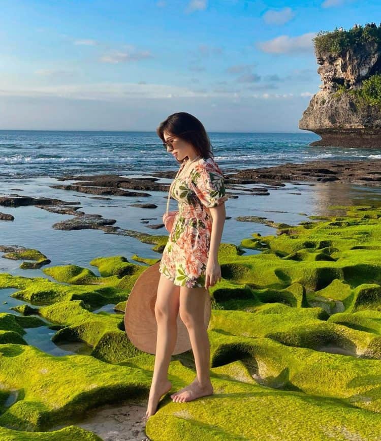 Wisatawan berdiri di atas batuan karang berwarna hijau tepi pantai